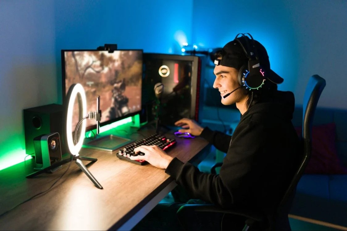 Gamer wearing headset at desk with dual monitors, ring light, and blue/green RGB lighting